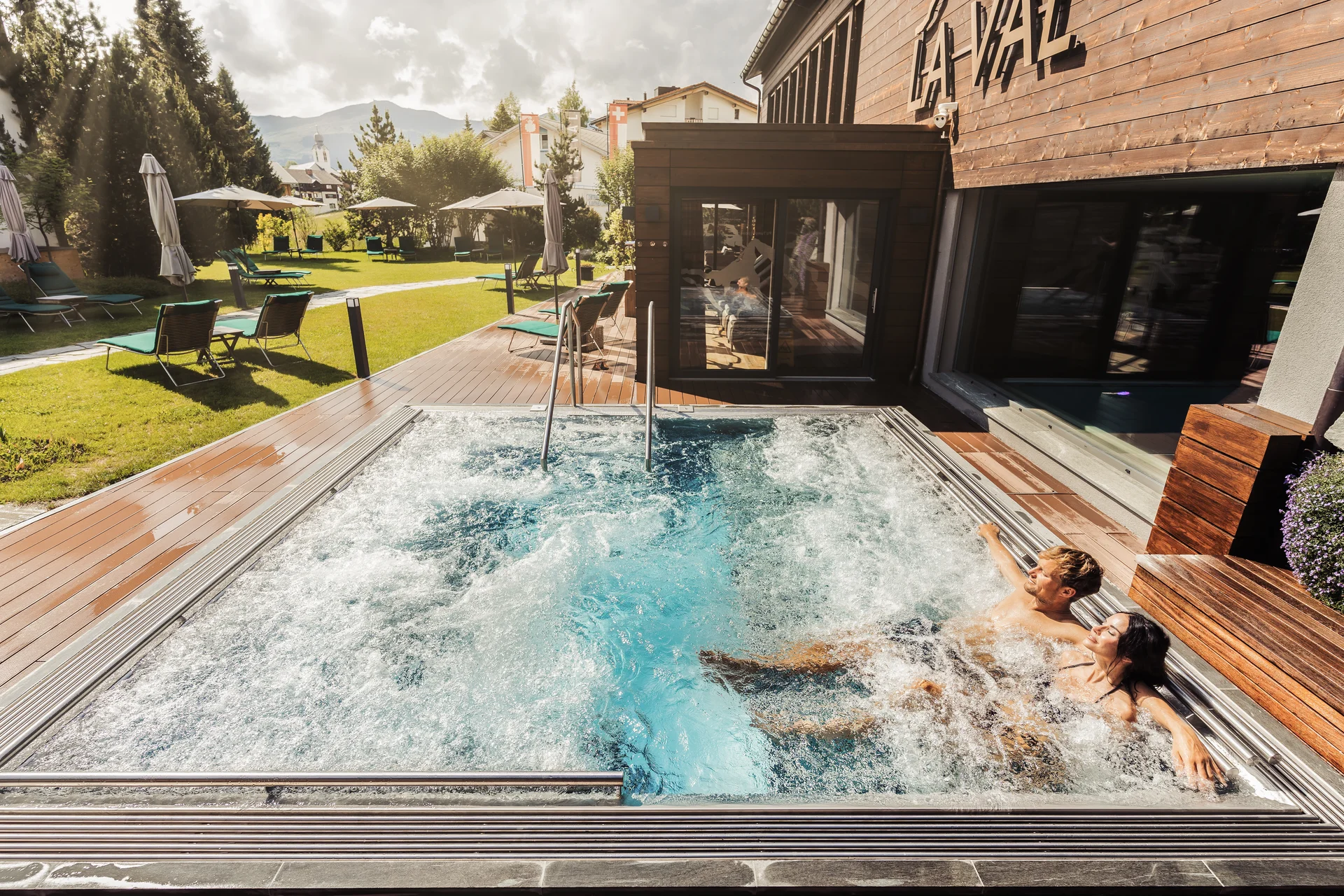 Paar entspannt im Whirlpool neben Hotel mit Garten und Liegestühlen
