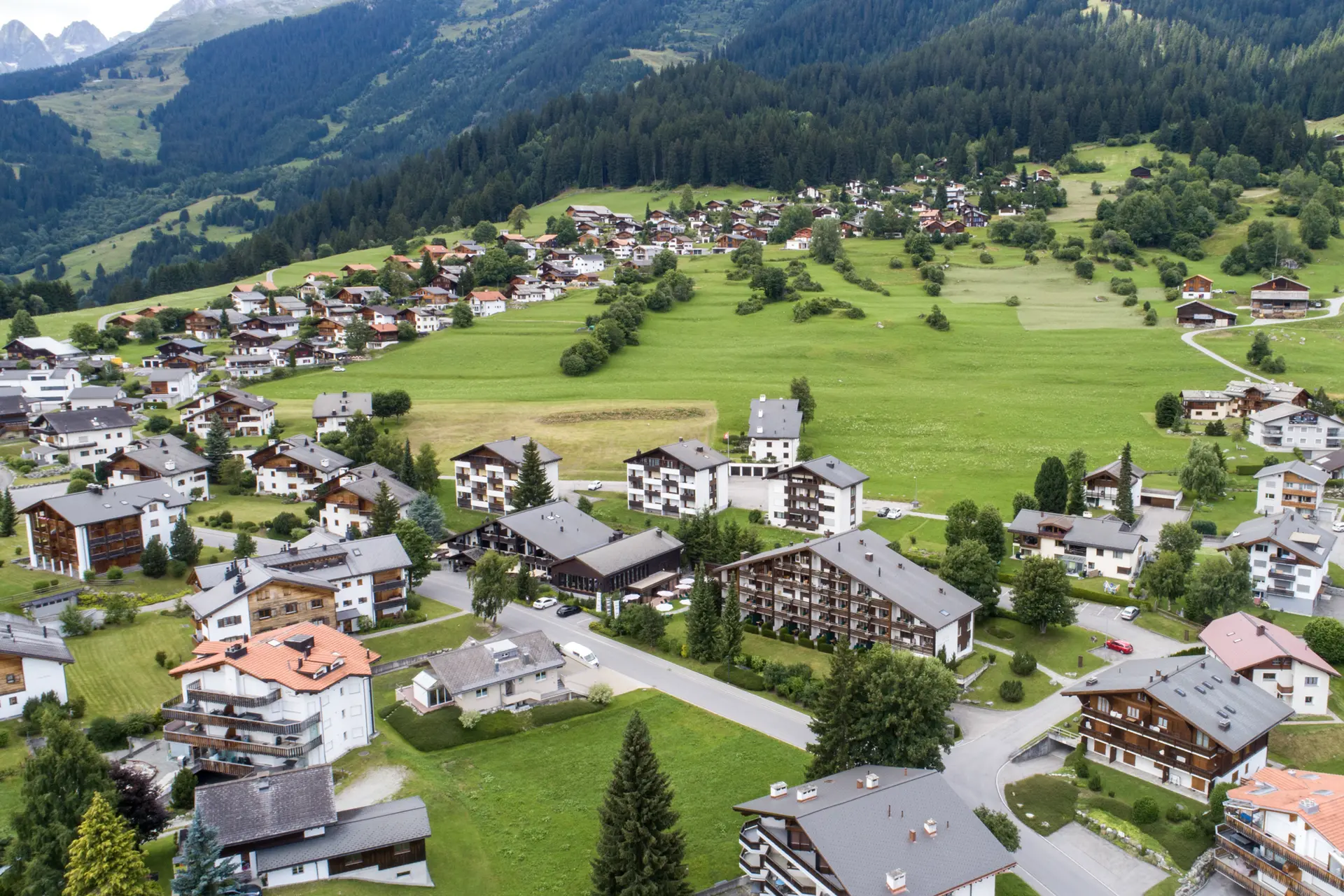 Eindrücke aus dem La Val Hotel & Spa Bergdorf mit Häusern, Wiesen und Wald am Fuß eines Berges