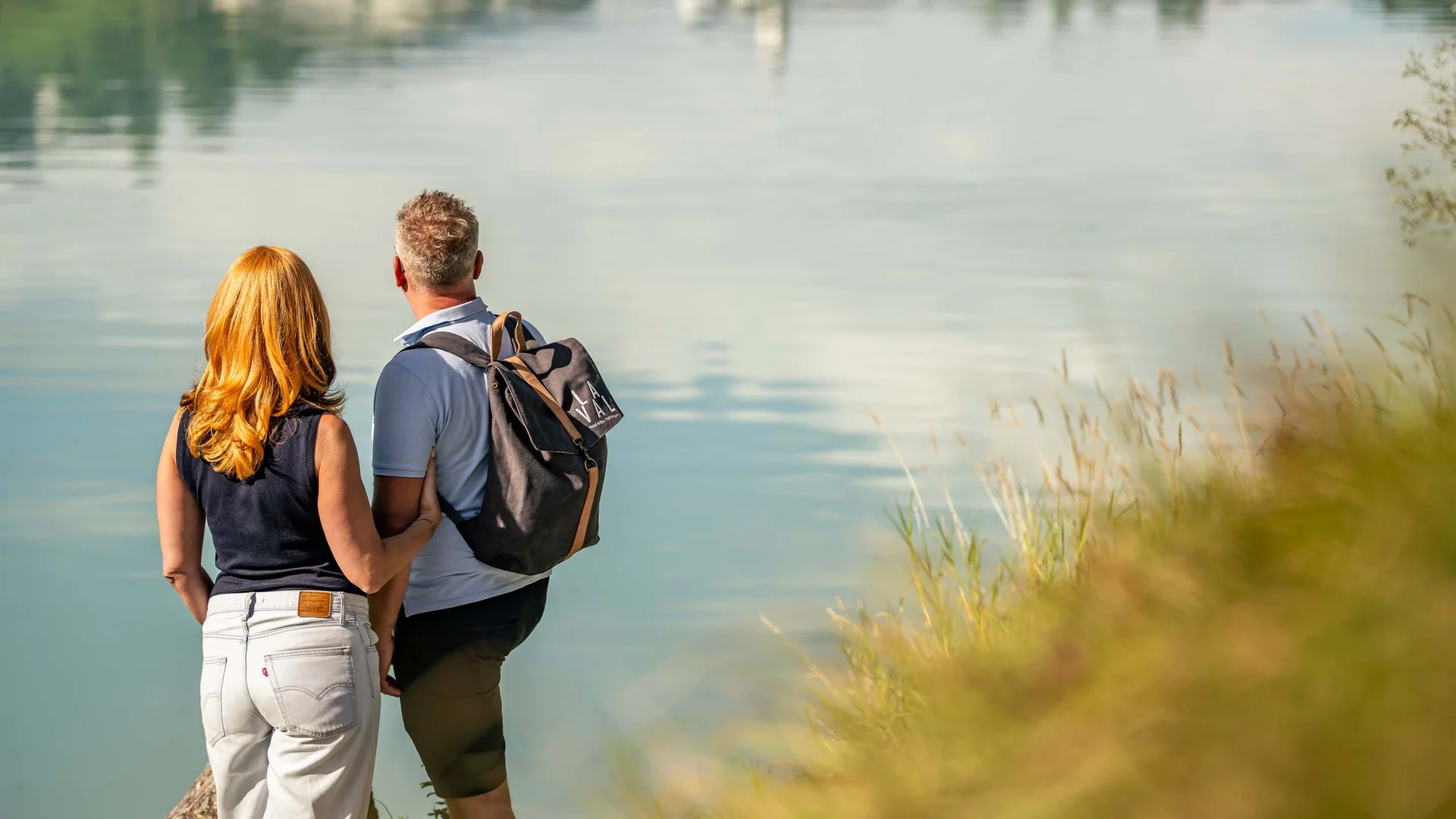 Paar steht am Ufer und blickt auf ruhigen See mit Spiegelung