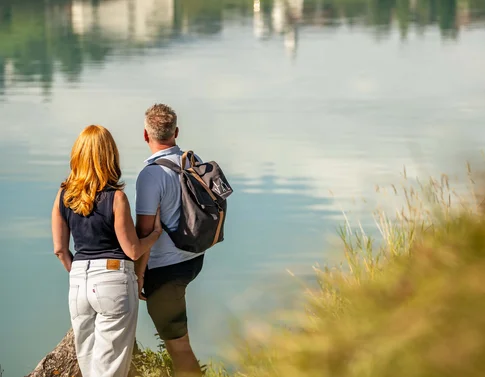 Paar steht am Ufer und blickt auf ruhigen See mit Spiegelung
