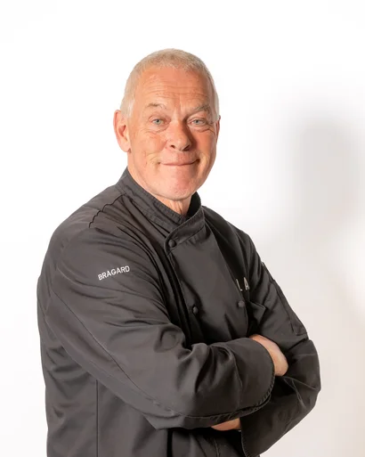 Älterer Koch in schwarzer Kochjacke vor weißem Hintergrund