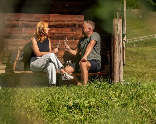 Storys aus dem La Val Hotel & Spa Paar sitzt auf Bank vor Holzhaus und stößt mit Weingläsern an