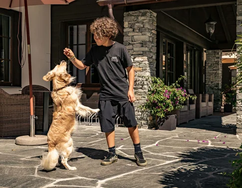 Wir sind Ihr Hundehotel in Graubünden! Kind füttert stehenden Hund vor Haus mit Steinmauer