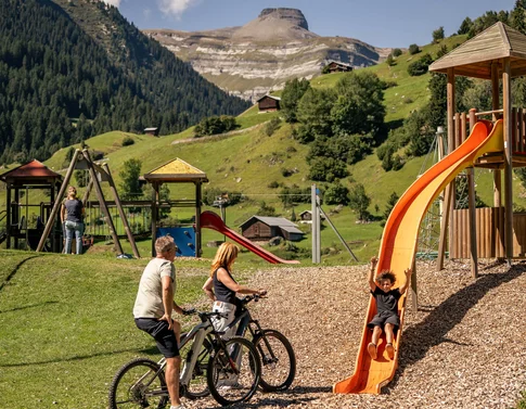 Kind rutscht auf Spielplatz, begleitet von Erwachsenen mit Fahrrädern in den Alpen