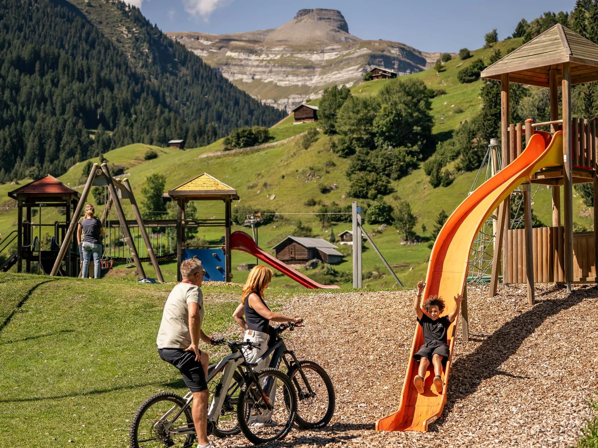 Kind rutscht auf Spielplatz, begleitet von Erwachsenen mit Fahrrädern in den Alpen