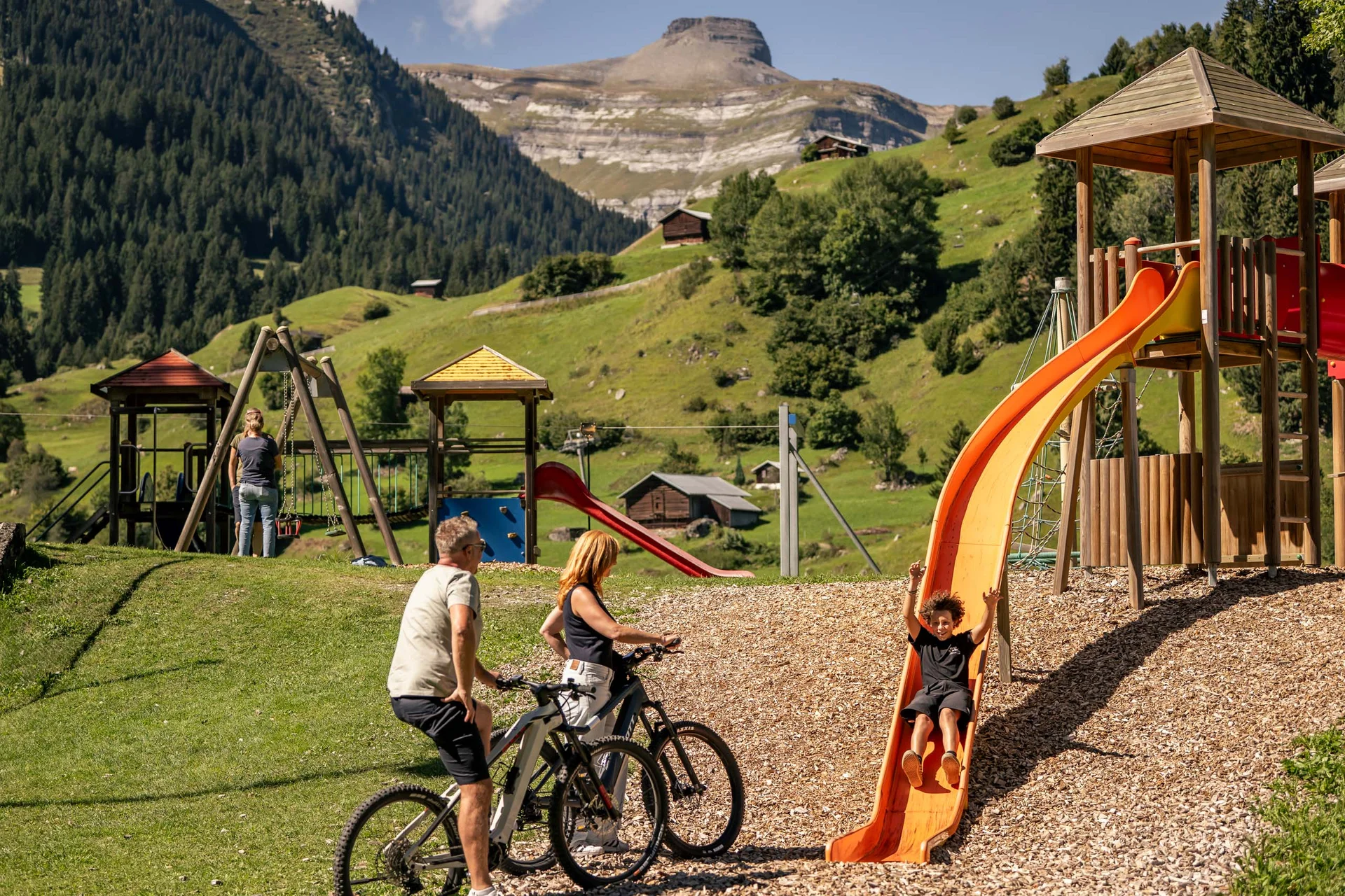 Kind rutscht auf Spielplatz, begleitet von Erwachsenen mit Fahrrädern in den Alpen