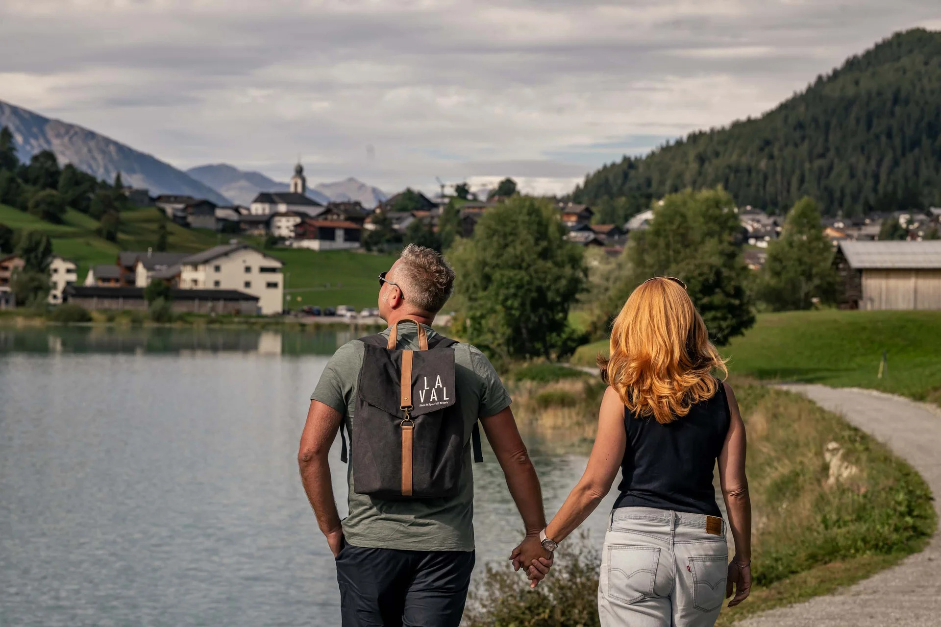 Paar hält Händchen beim Spaziergang an einem See mit Bergen und Dorf im Hintergrund