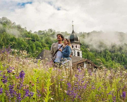 Couple standing in wildflower meadow near church in foggy mountain valley