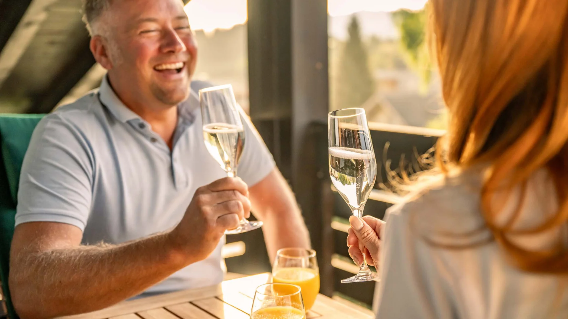 Paar stößt mit Champagnergläsern auf einem Balkon an, Orangensaftgläser stehen auf dem Tisch