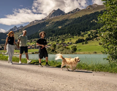 Ab ins Wellnesshotel – mit dem Hund in die Schweiz Familie spaziert mit Hund auf Weg neben See in den Bergen