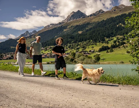 Wir sind Ihr Hundehotel in Graubünden! Familie spaziert mit Hund auf Weg neben See in den Bergen