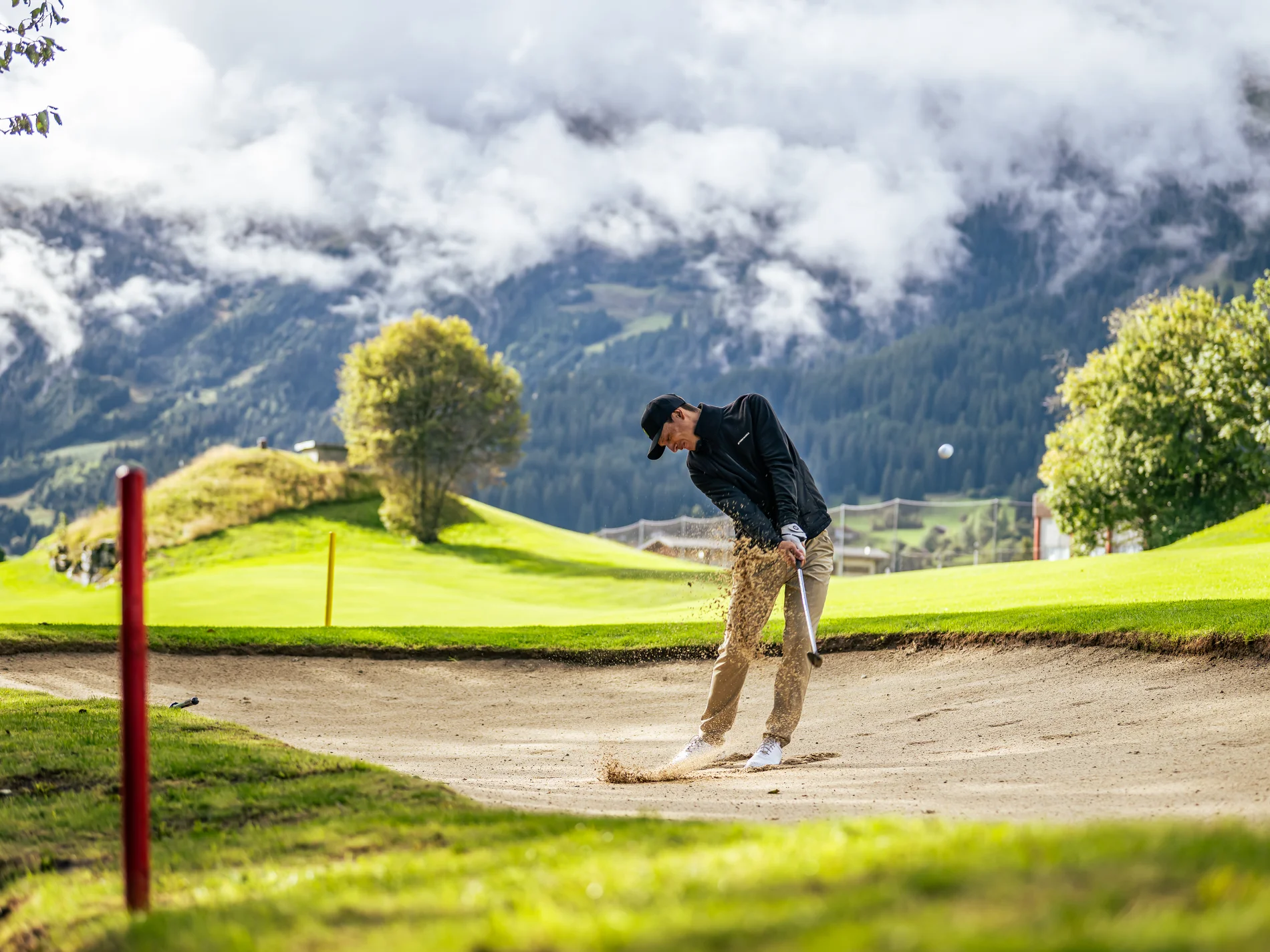 Golfer schlägt Ball aus dem Sandbunker auf grünem Golfplatz vor Bergkulisse