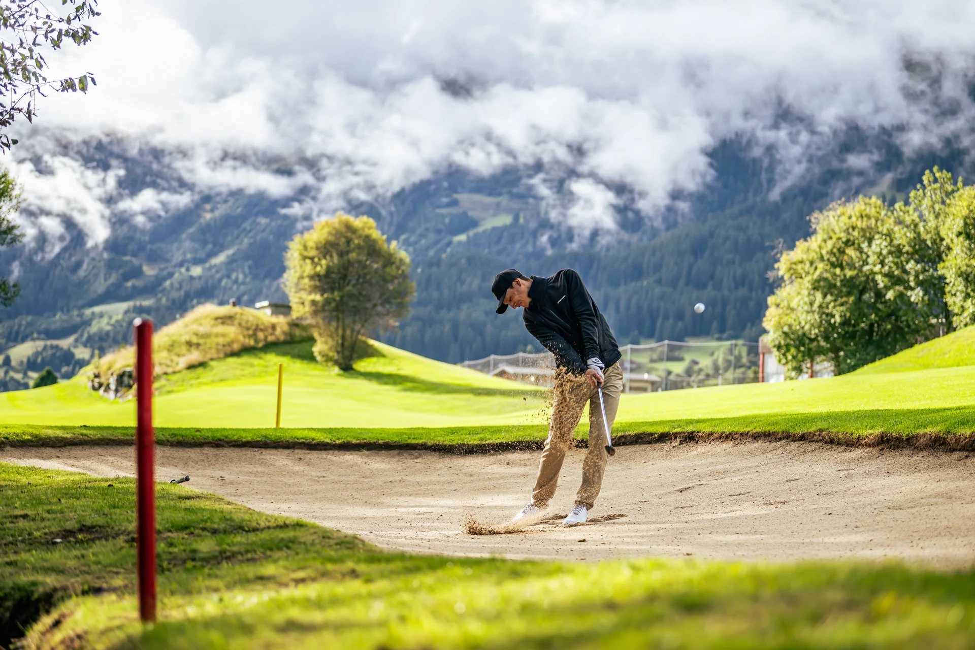 Golfer schlägt Ball aus dem Sandbunker auf grünem Golfplatz vor Bergkulisse