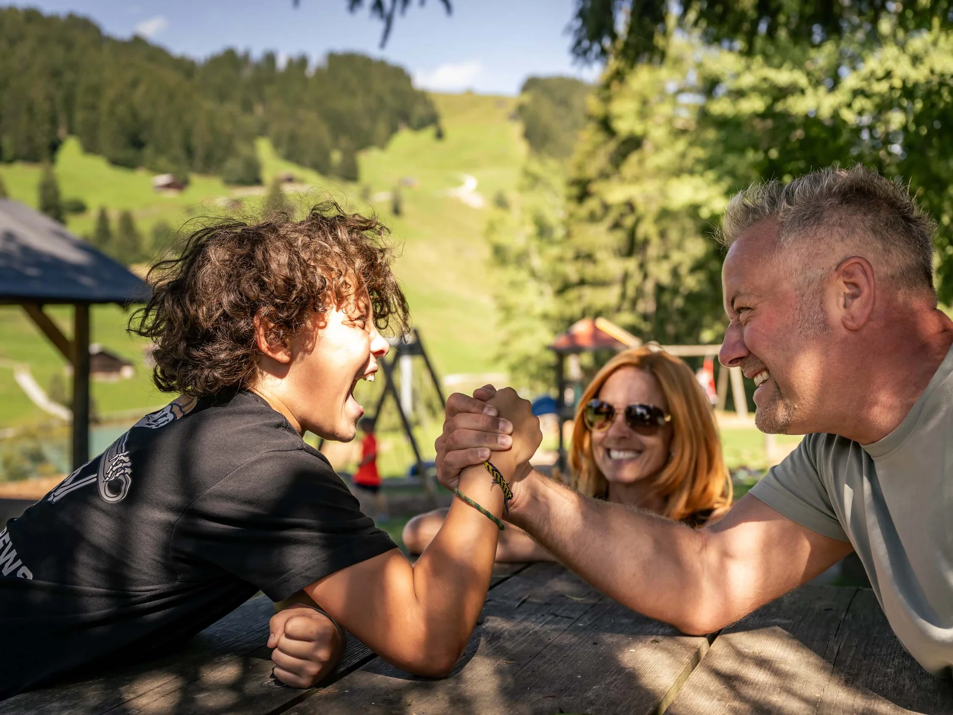 Junge und Mann beim Armdrücken an einem Picknicktisch, Frau lächelt im Hintergrund