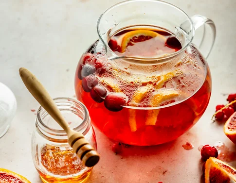 Glaskrug mit fruchtigem Heißgetränk, Honigglas mit Honiglöffel und frische Orangenscheiben