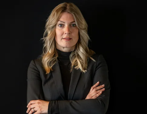 Frau mit blonden Haaren in schwarzem Anzug vor dunklem Hintergrund, Arme verschränkt