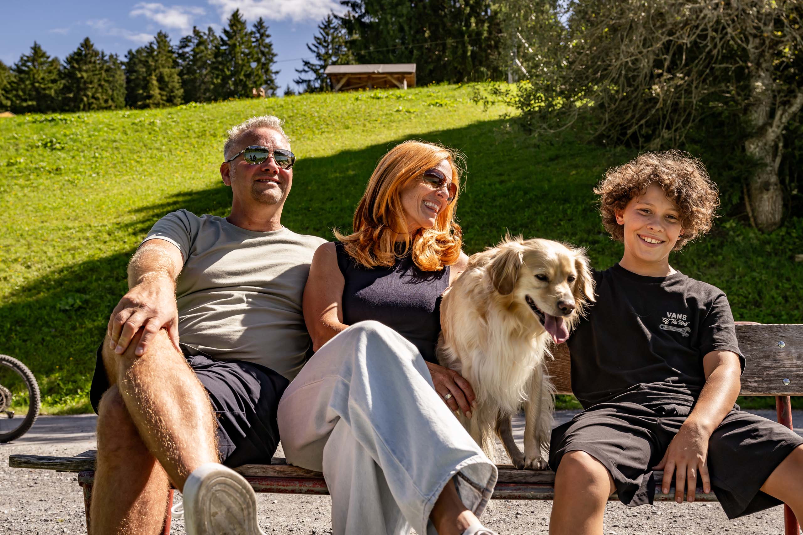 Wir sind Ihr Hundehotel in Graubünden! Familie mit Hund sitzt auf Parkbank bei sonnigem Wetter und grünem Hügel